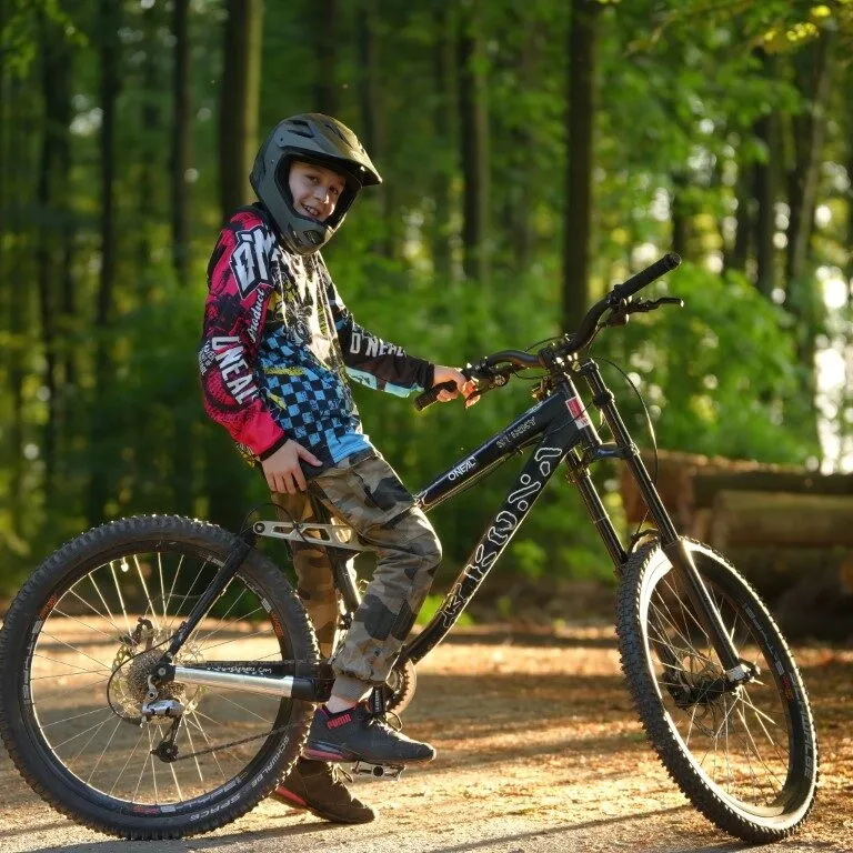 Kind in MTB-Ausrüstung mit Fullface-Helm auf Downhill-Bike im Wald